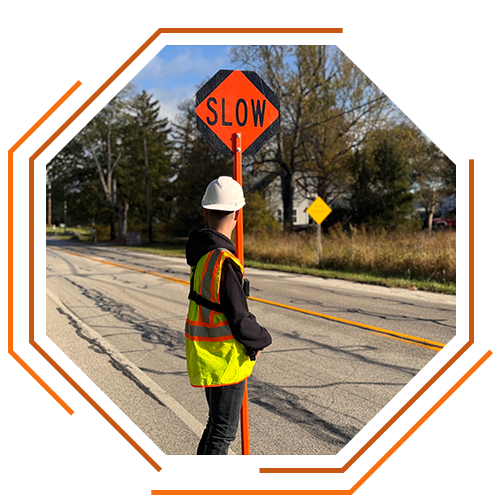 stylized photo: a KSK employee in yellow and orange safety gear and a white hardhat holds an orange "SLOW" sign for traffic management